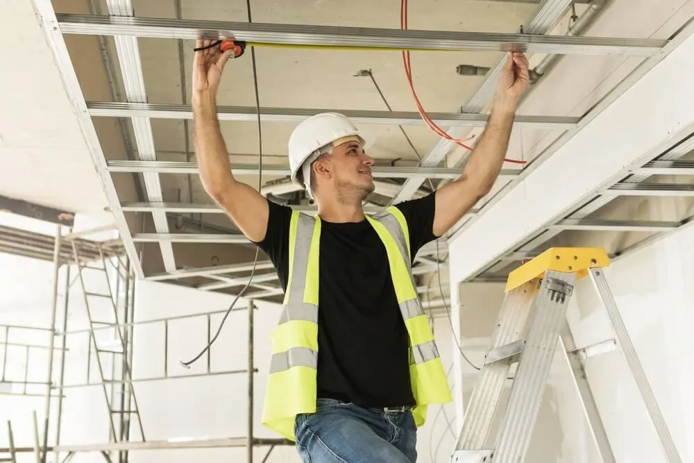Tradesman in a hard hat and hi-vis vest measuring a ceiling frame from a stepladder