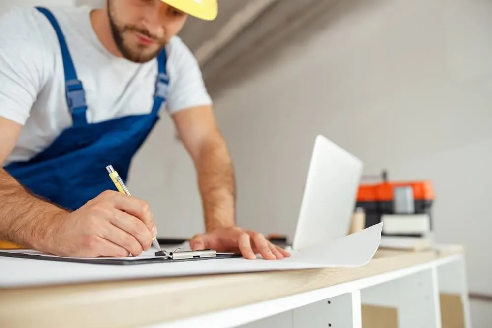 Builder in a hard hat and overalls writing notes at a workbench beside a laptop