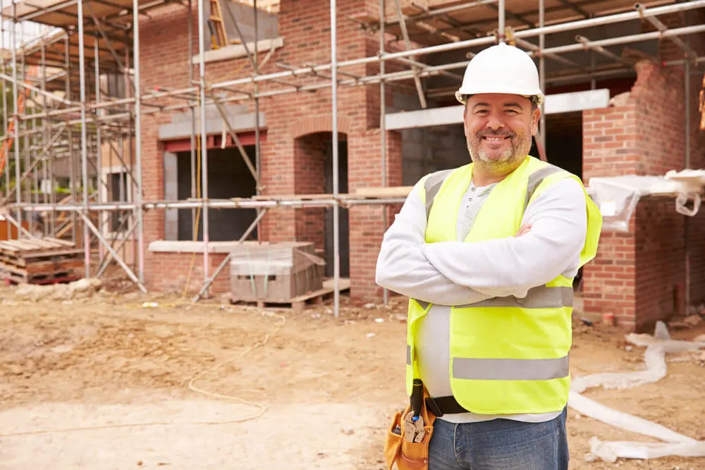Smiling builder in a hard hat and hi-vis vest with arms folded on a building site